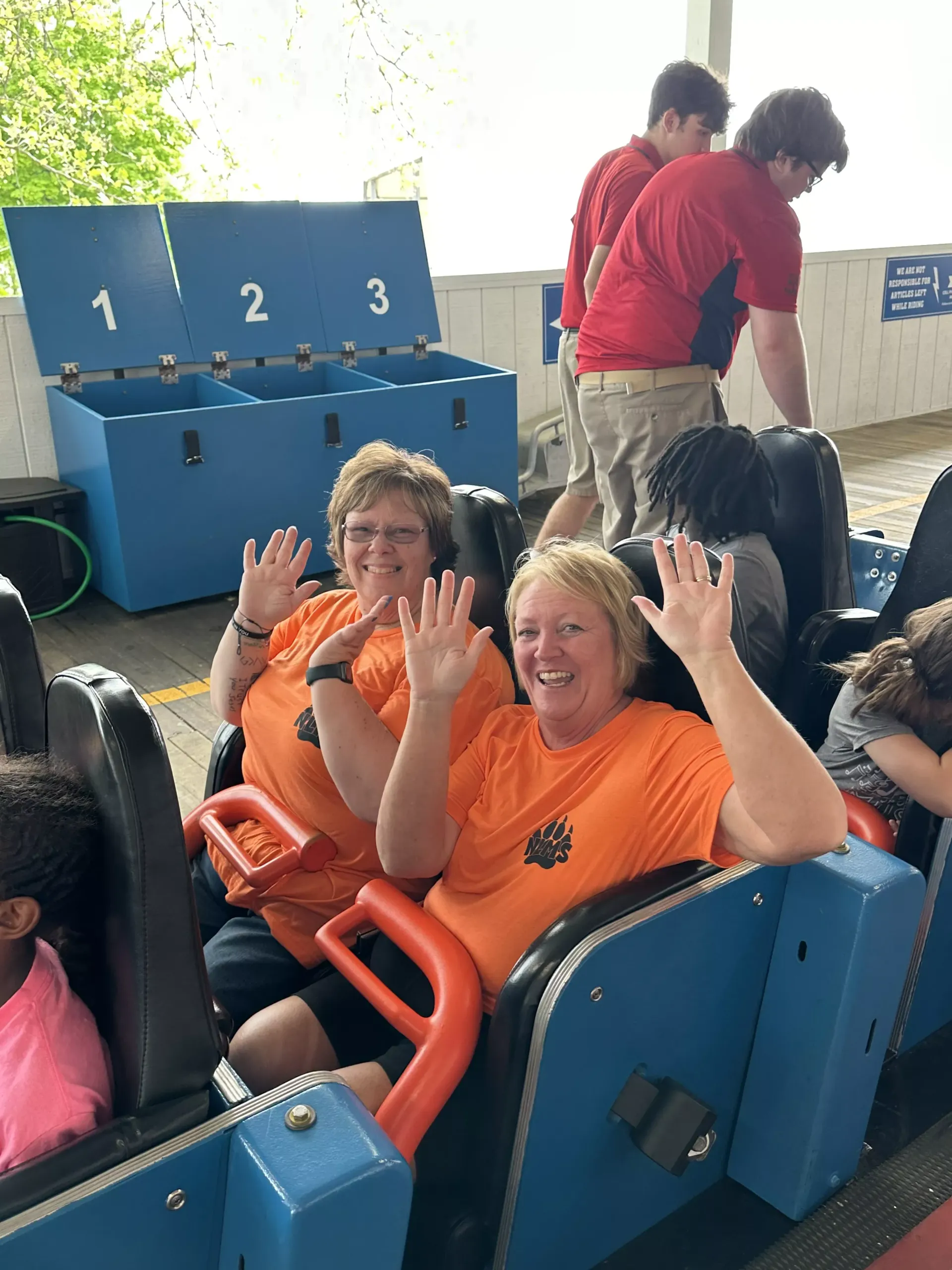 Two people in orange shirts waving from a blue amusement ride car with numbered seats