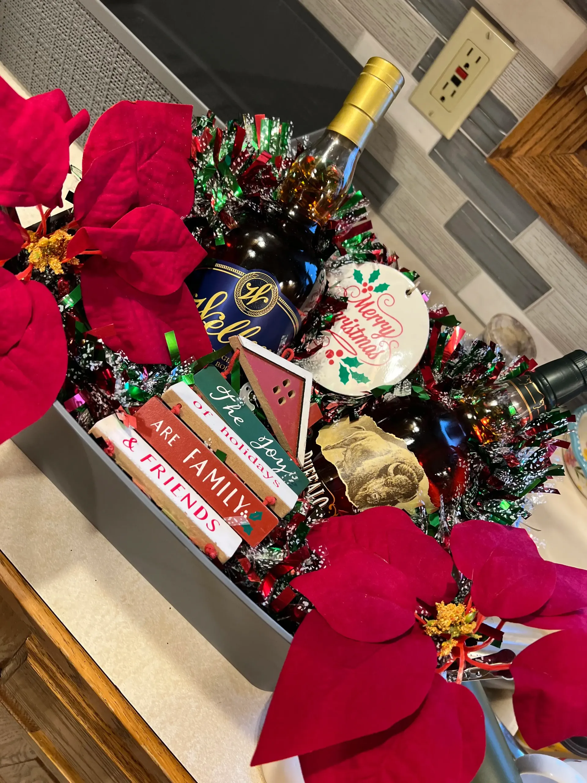 Holiday decorated bottle basket with red poinsettias, tinsel, and gift tags on a countertop.