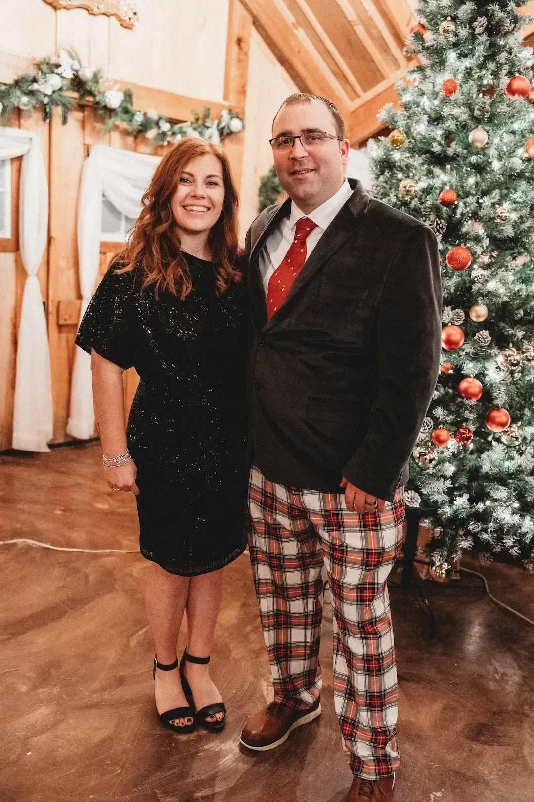 Two people posing indoors beside a decorated Christmas tree, wearing festive formal outfits.
