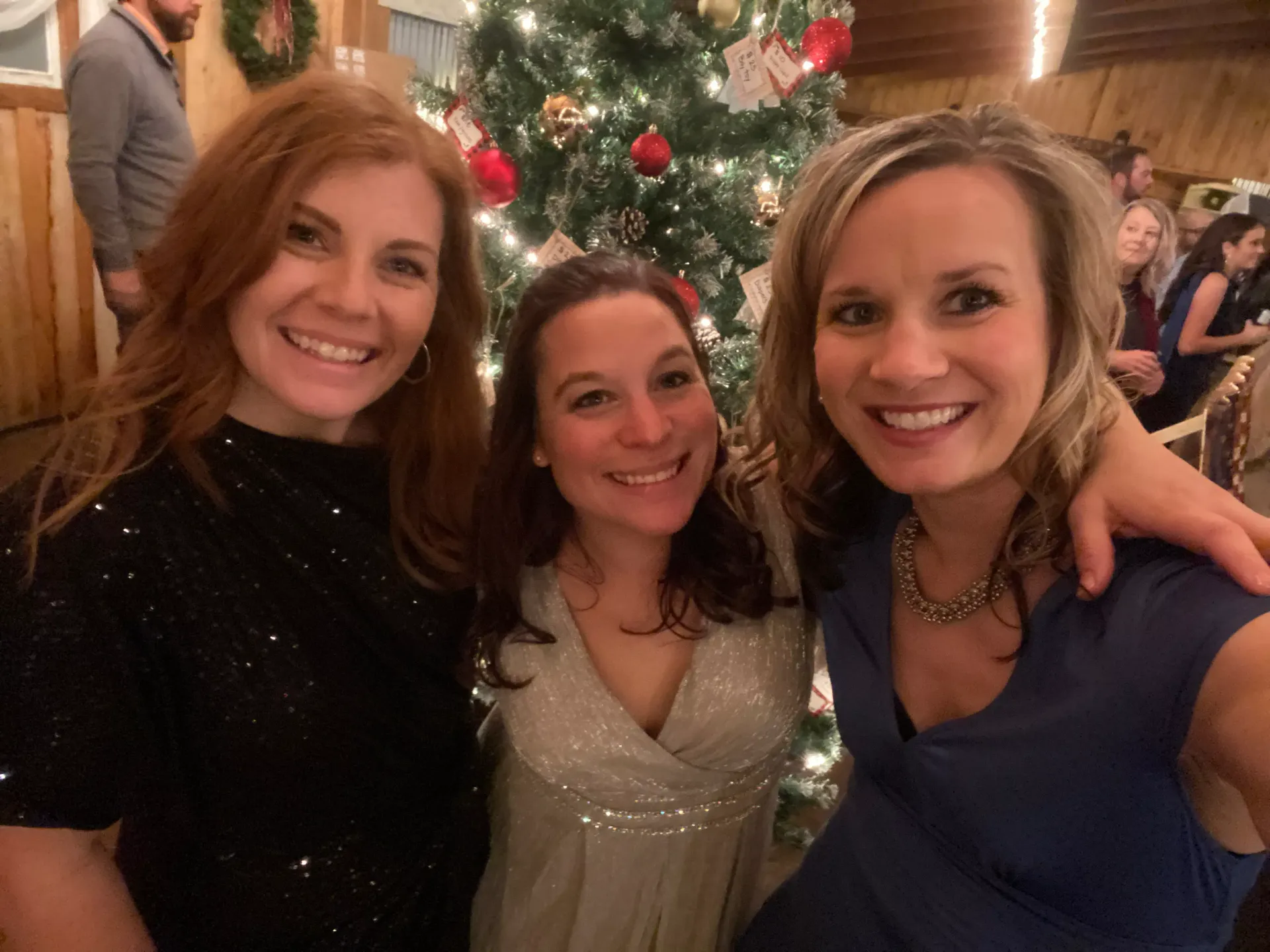 Three smiling people posing in front of a decorated Christmas tree at a festive indoor gathering