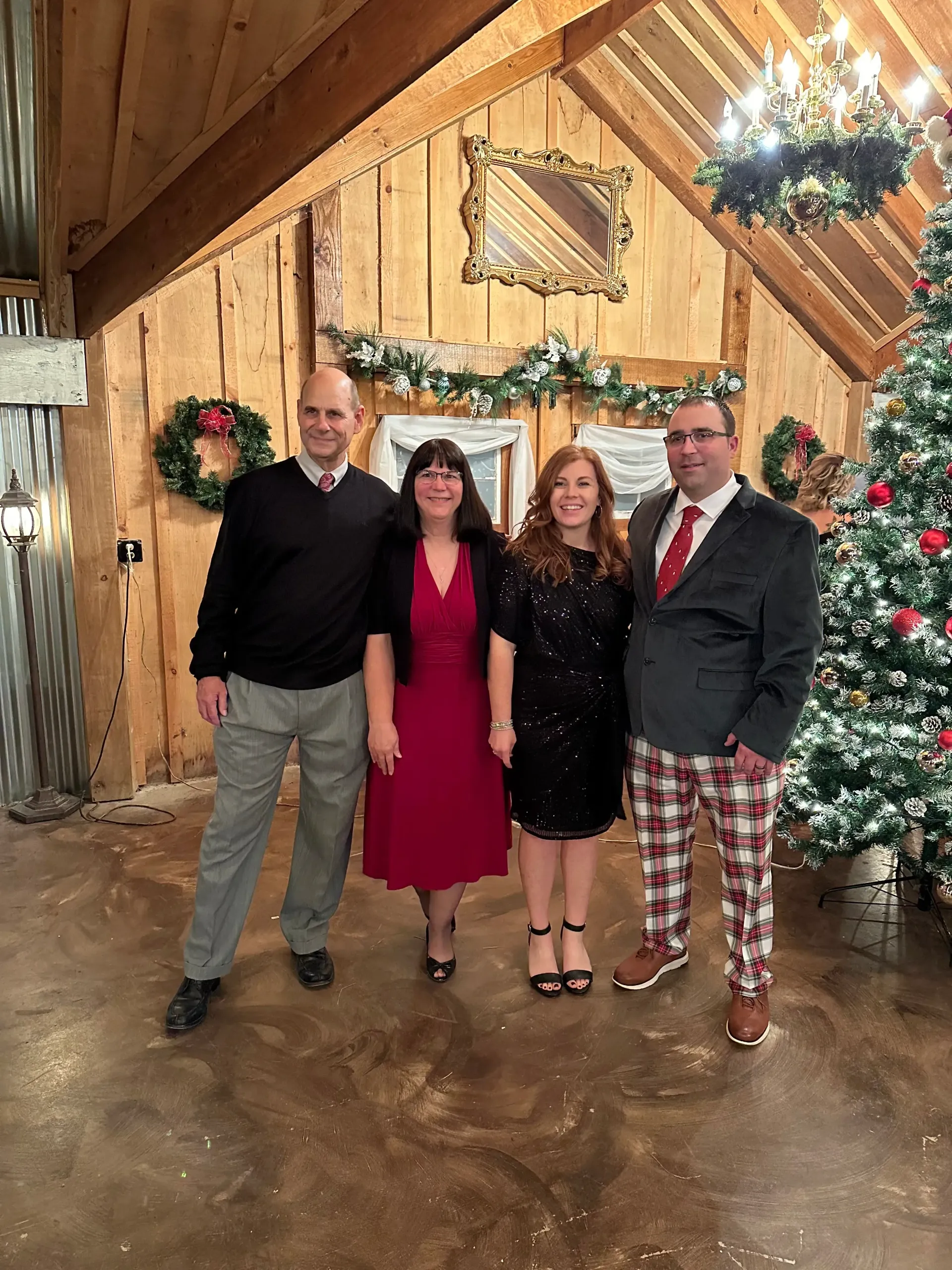 Four people posing in festive holiday attire in a rustic, decorated room with a Christmas tree.