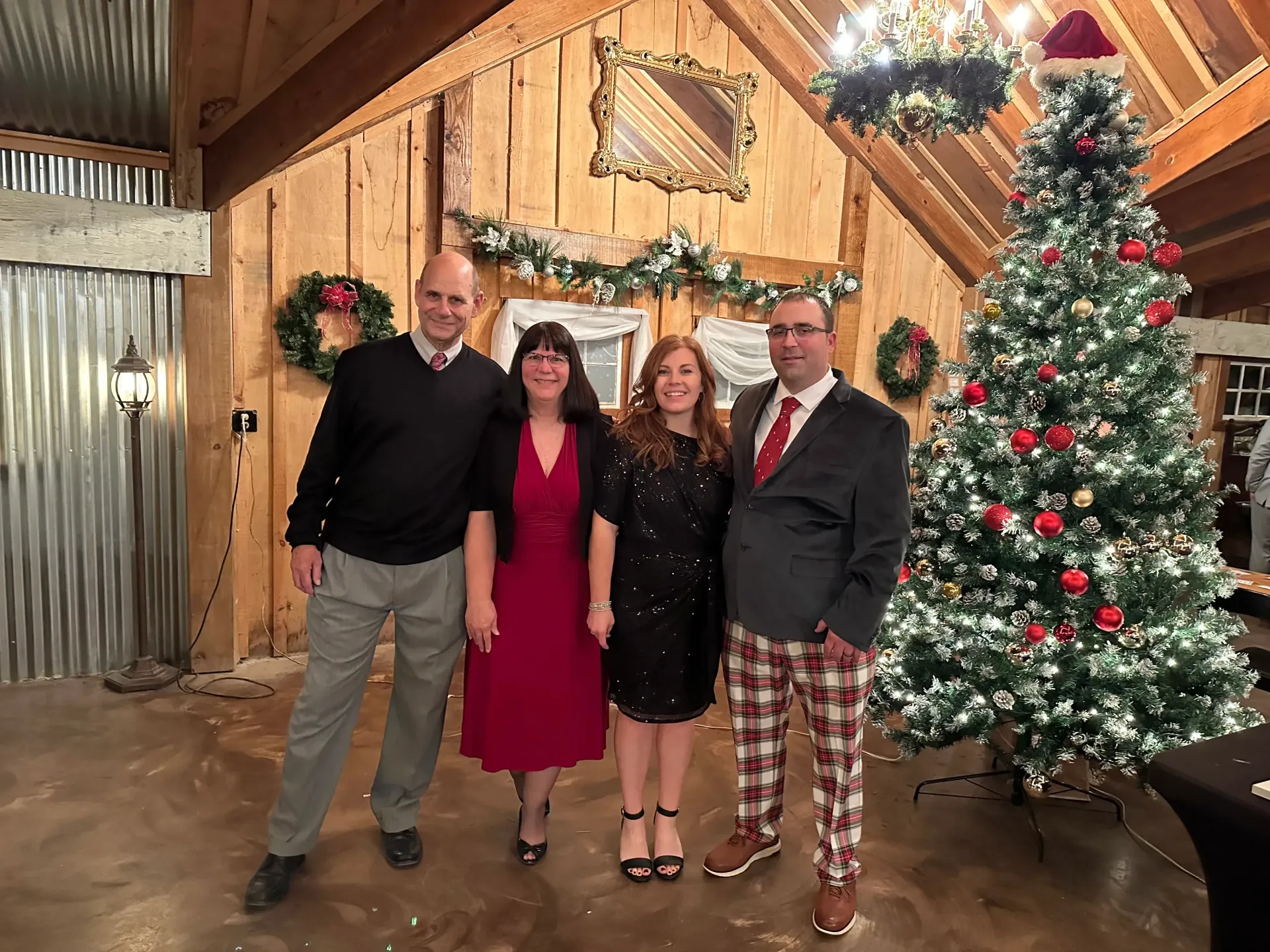 Four people posing indoors beside a decorated Christmas tree and rustic wood wall.