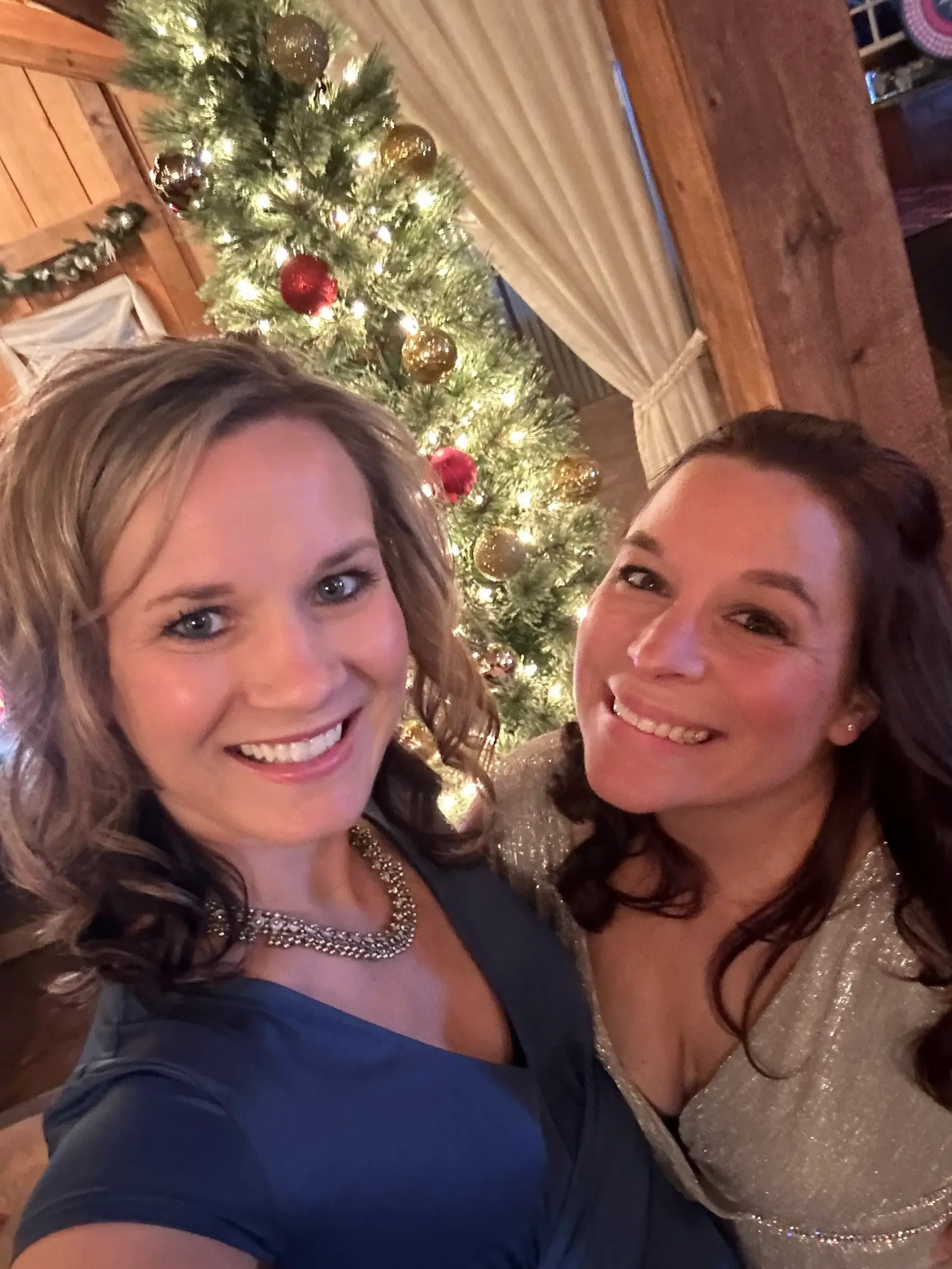 Two women smiling for a selfie by a lit Christmas tree indoors
