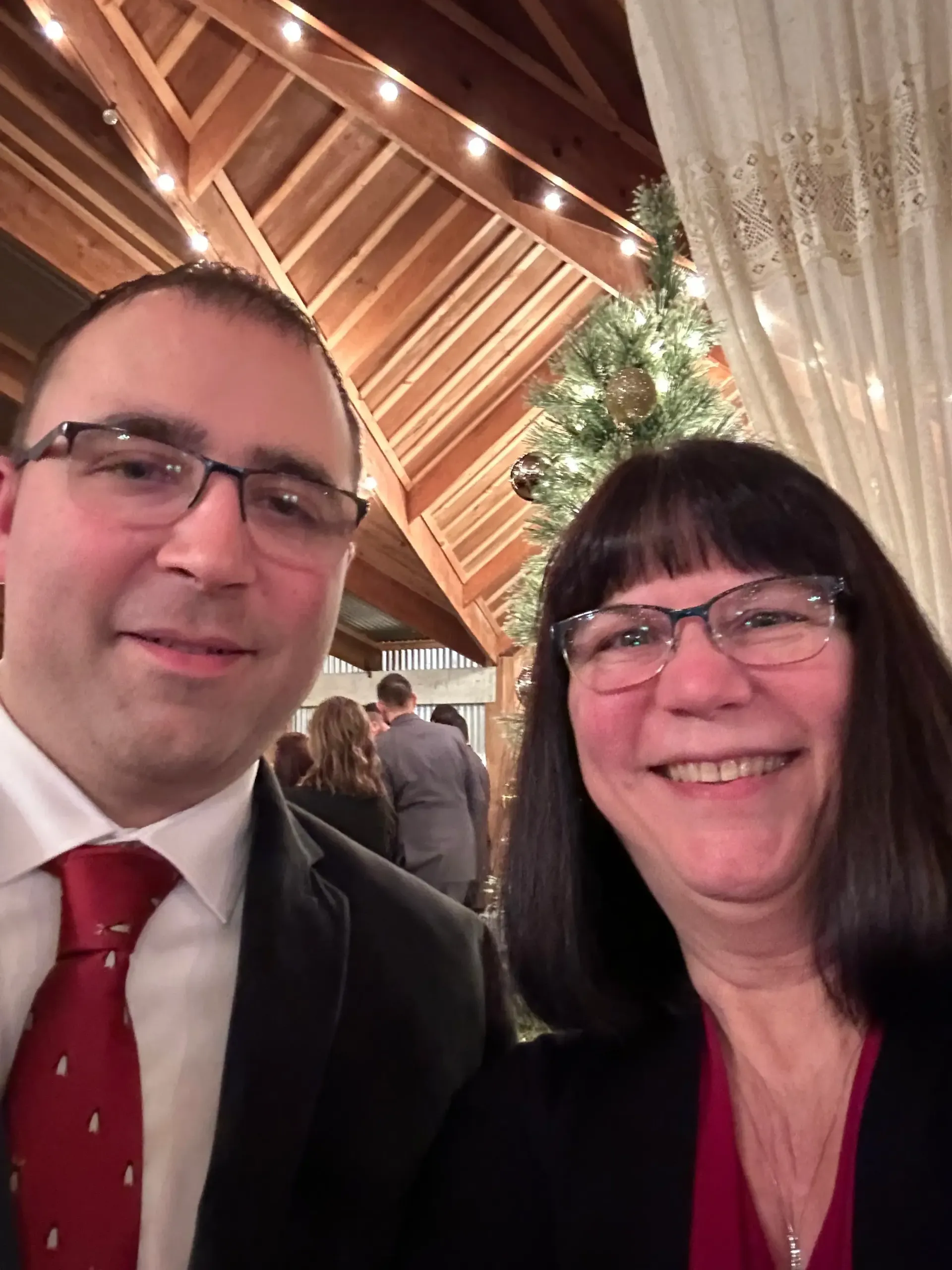 Two smiling people in formal attire at an indoor event with warm wood ceiling and string lights
