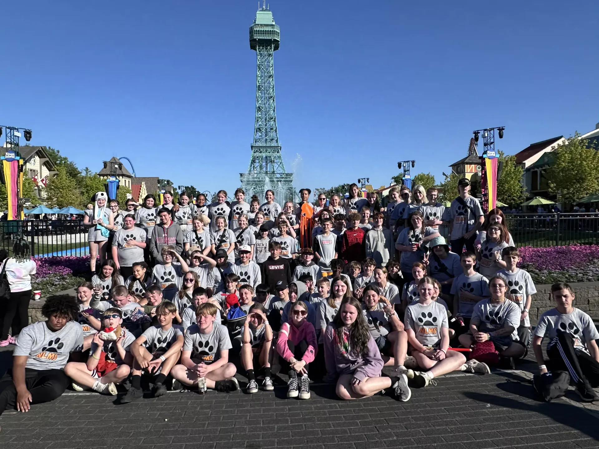 Large group posing in front of a tall tower on a sunny day, with colorful flower beds and blue sky.