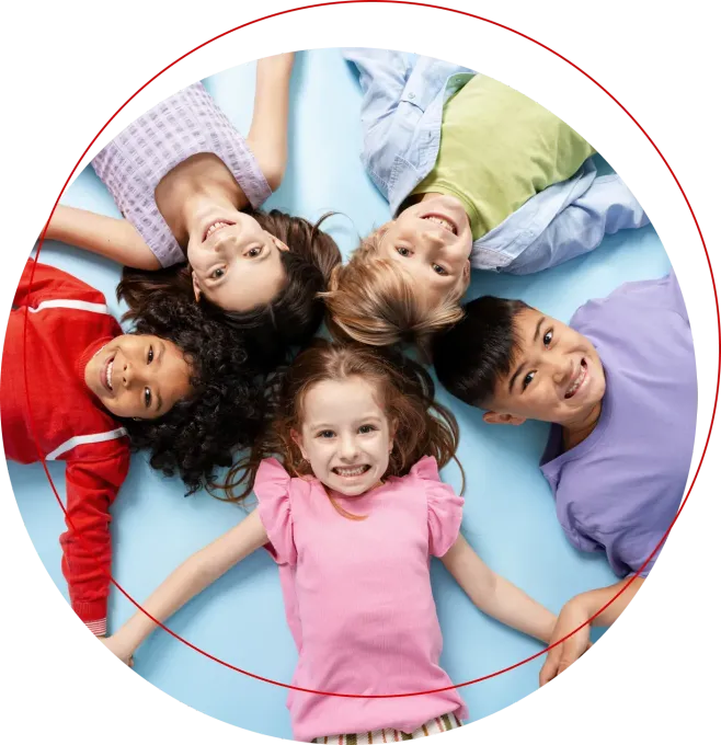 Five smiling children lying in a circle on the floor, heads together, looking up at the camera.