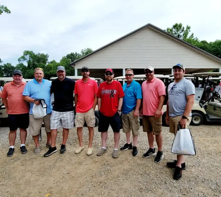 Eight men stand together on a gravel lot in front of a beige building, posing for a group photo.