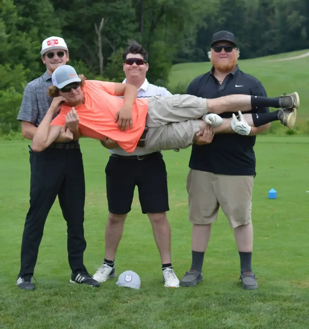 Four golfers on a course carry a man horizontally between them, all wearing sunglasses.