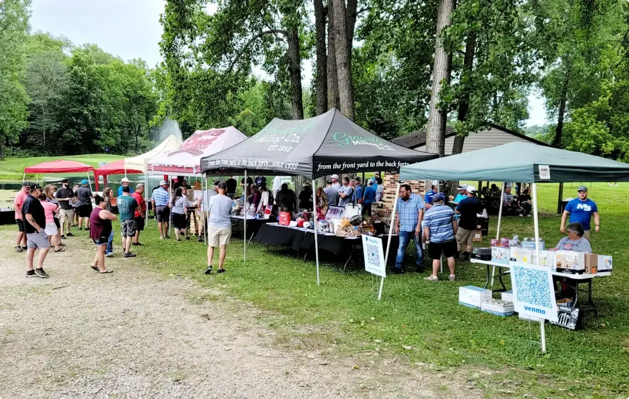 Outdoor community market with tents, tables, and people browsing in a wooded park