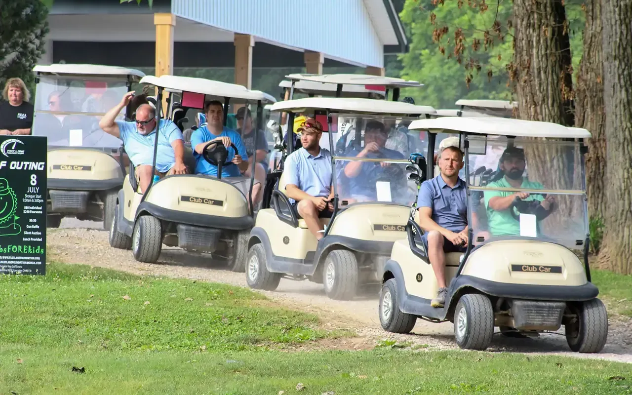 Golf carts lined up with passengers on a grassy path beside a building.