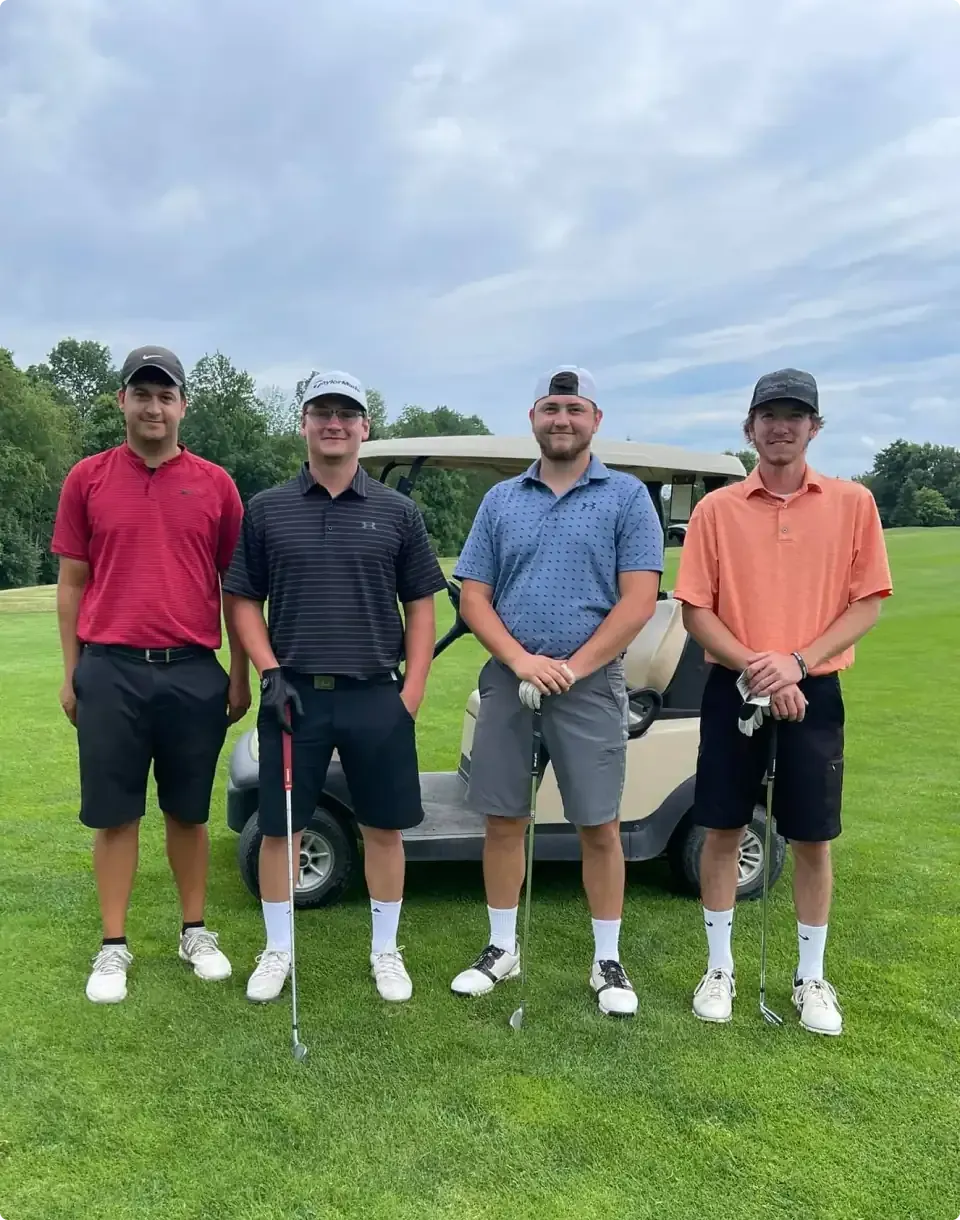 Four golfers standing on a green fairway, holding clubs, with trees and a cloudy sky behind them.