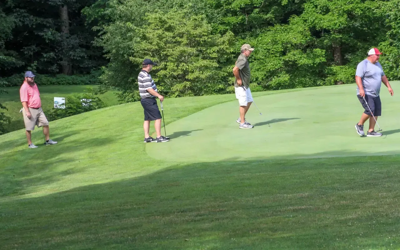 Four golfers standing on a green fairway near the woods, waiting with clubs.