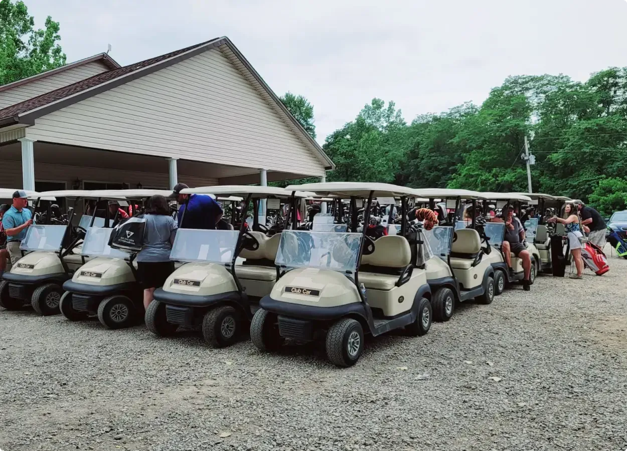 Row of golf carts parked outside a pavilion with people gathered nearby on a gravel lot