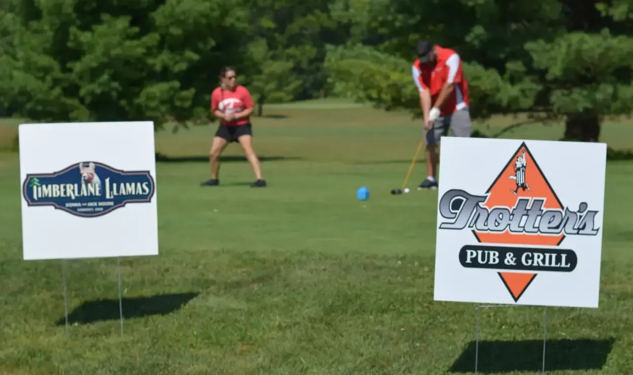 Two golfers on a green field behind sponsor signs for a putting contest.