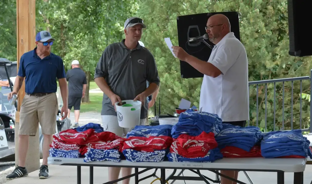Three men at an outdoor table with red and blue merchandise, one holding papers under a canopy.
