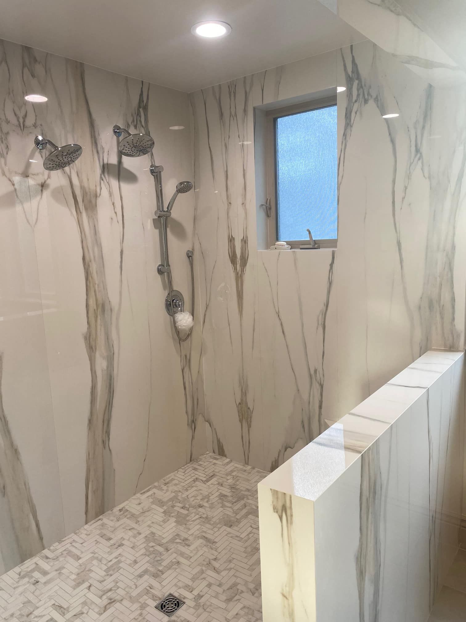 Modern marble-tiled shower with multiple showerheads, pebble floor, and small frosted window