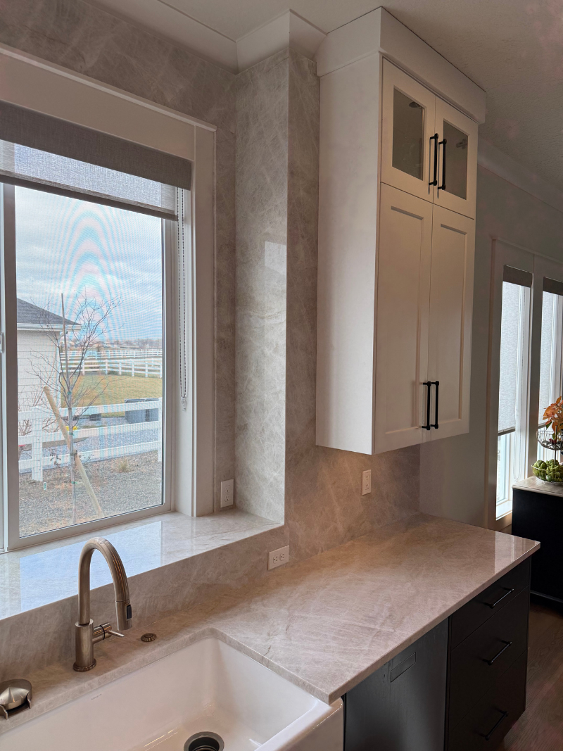 Bright kitchen with large window, stone backsplash, white cabinets, and countertop sink.