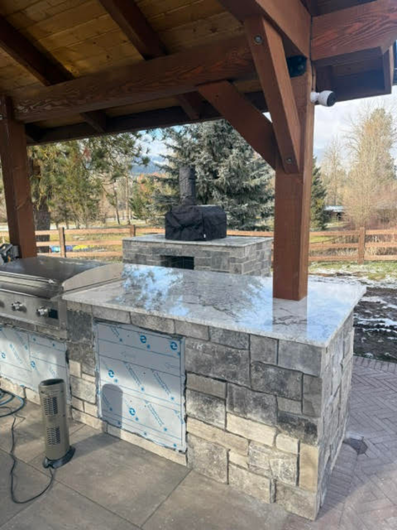 Stone outdoor grill under a wooden pavilion with a black smoker on top and a park in the background