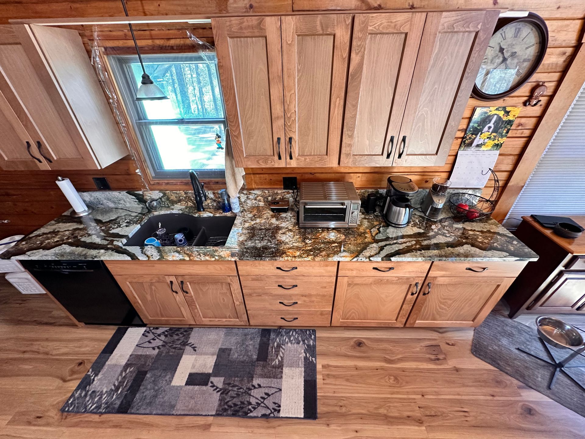 Wooden kitchen with granite counters, sink under a window, and a black rug on the hardwood floor.