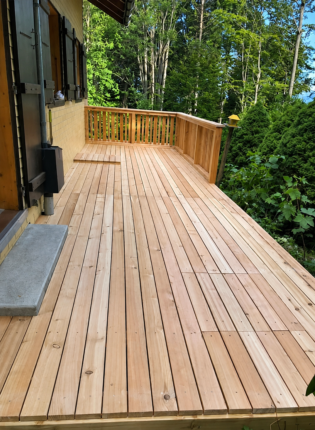 Terrasse en bois avec rambarde jouxtant une maison, bordée d'arbres et d'arbustes verdoyants.