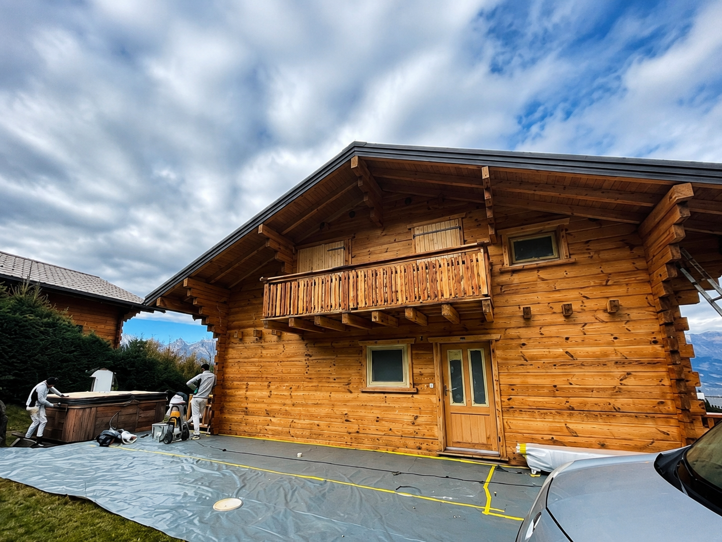 Chalet en bois avec balcon et murs en rondins sculptés, sous un ciel nuageux, dans une cour pavée avec des voitures garées