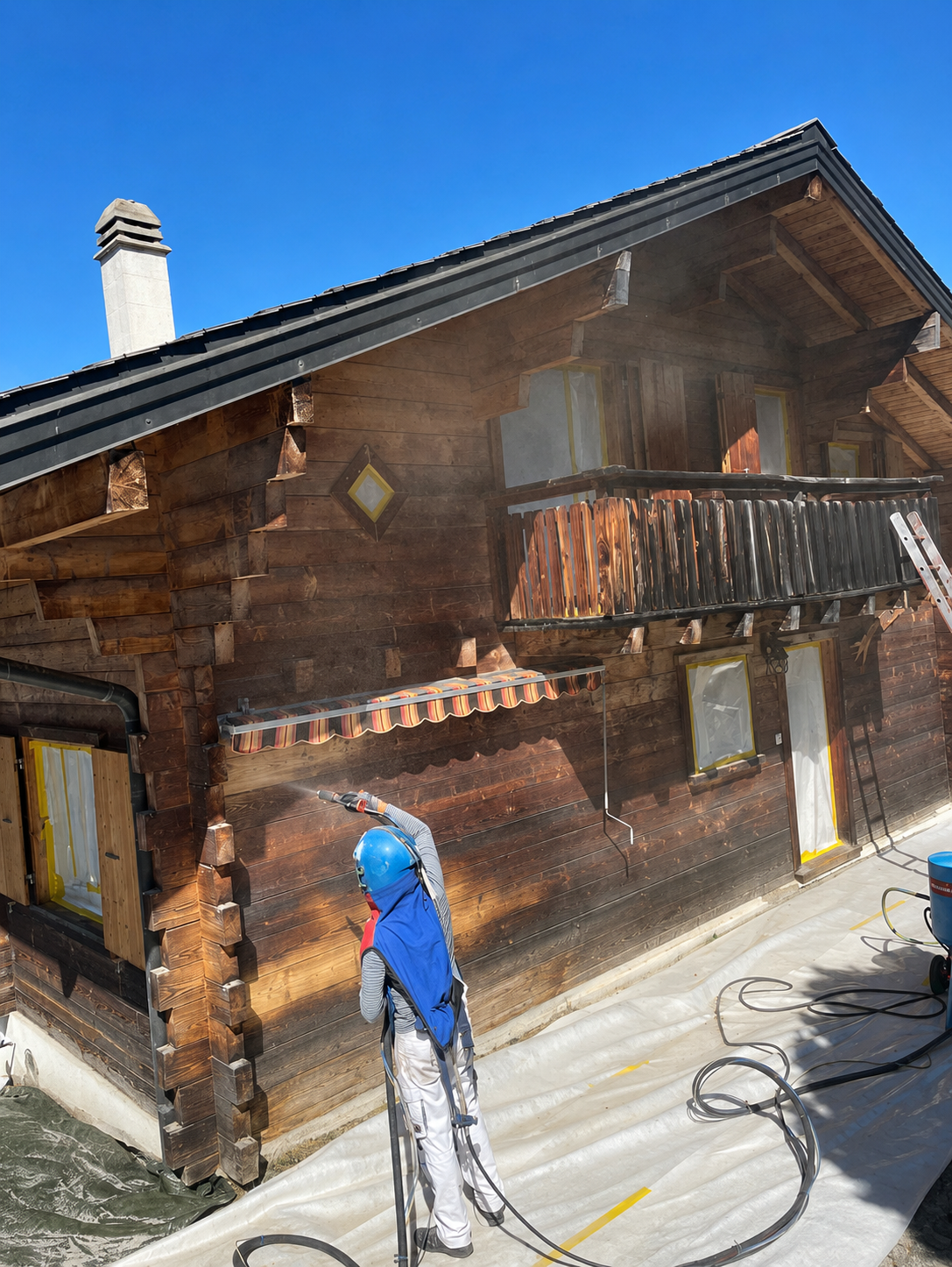 Un ouvrier nettoie au nettoyeur haute pression une maison de style chalet en bois avec balcon par une journée ensoleillée