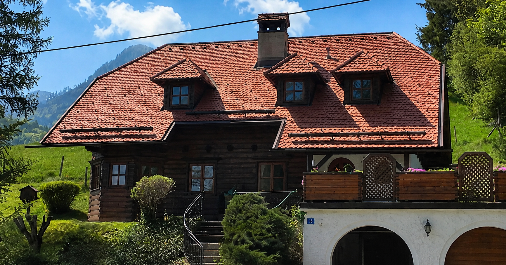 Maison rustique avec un toit de tuiles rouges, des lucarnes et une cheminée en pierre dans un cadre verdoyant.
