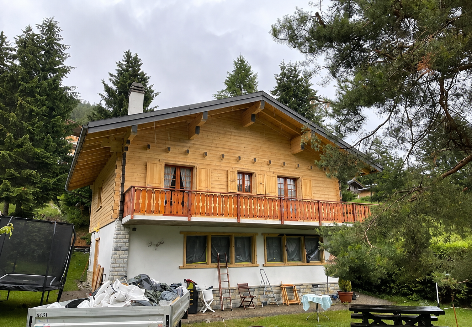 Chalet à deux étages avec bardage en bois jaune, balcon orange et niveau inférieur blanc entouré d'arbres.