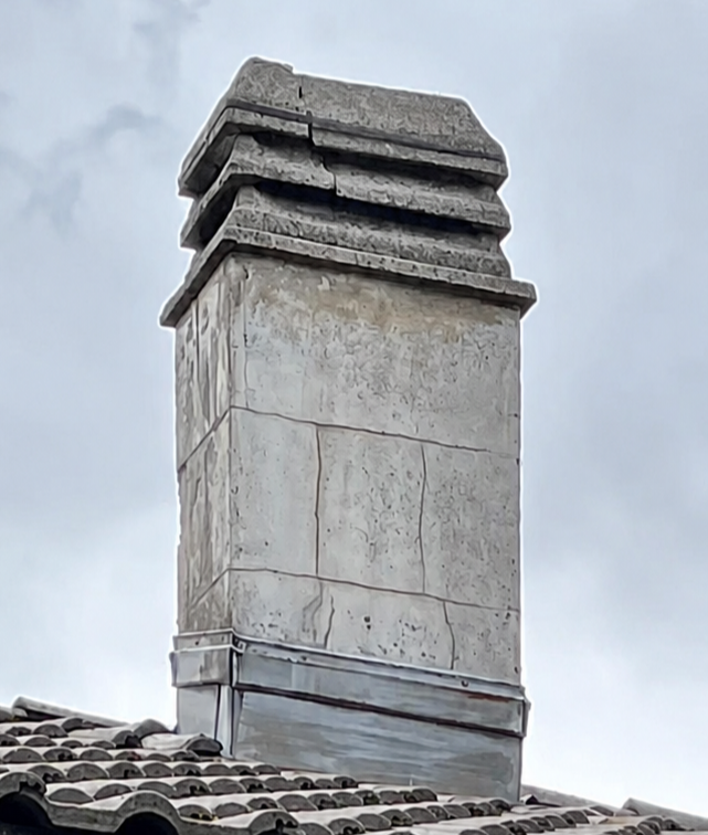 Cheminée en pierre au chapeau usé par le temps, sur un toit de tuiles se détachant sur un ciel nuageux
