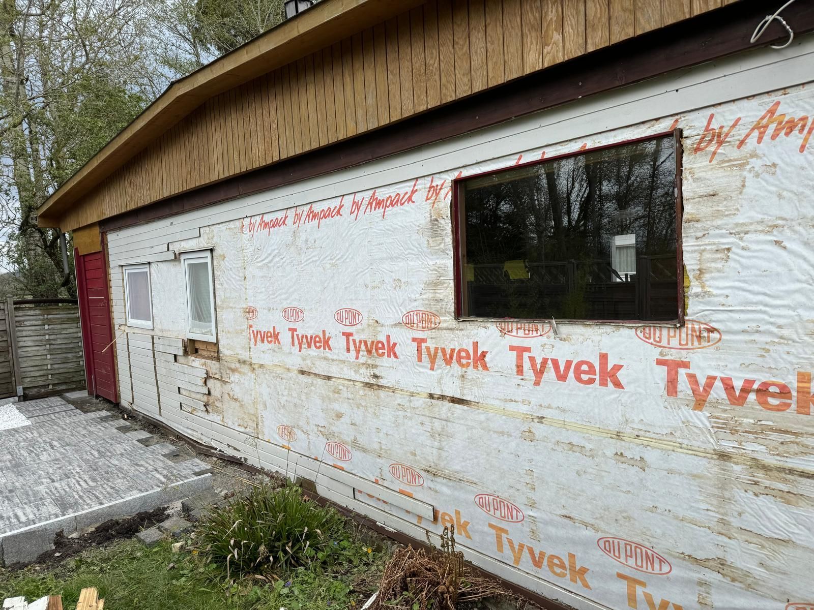 Maison avec façade recouverte de Tyvek, fenêtres, porte rouge et bardage inachevé sous un toit en pente.