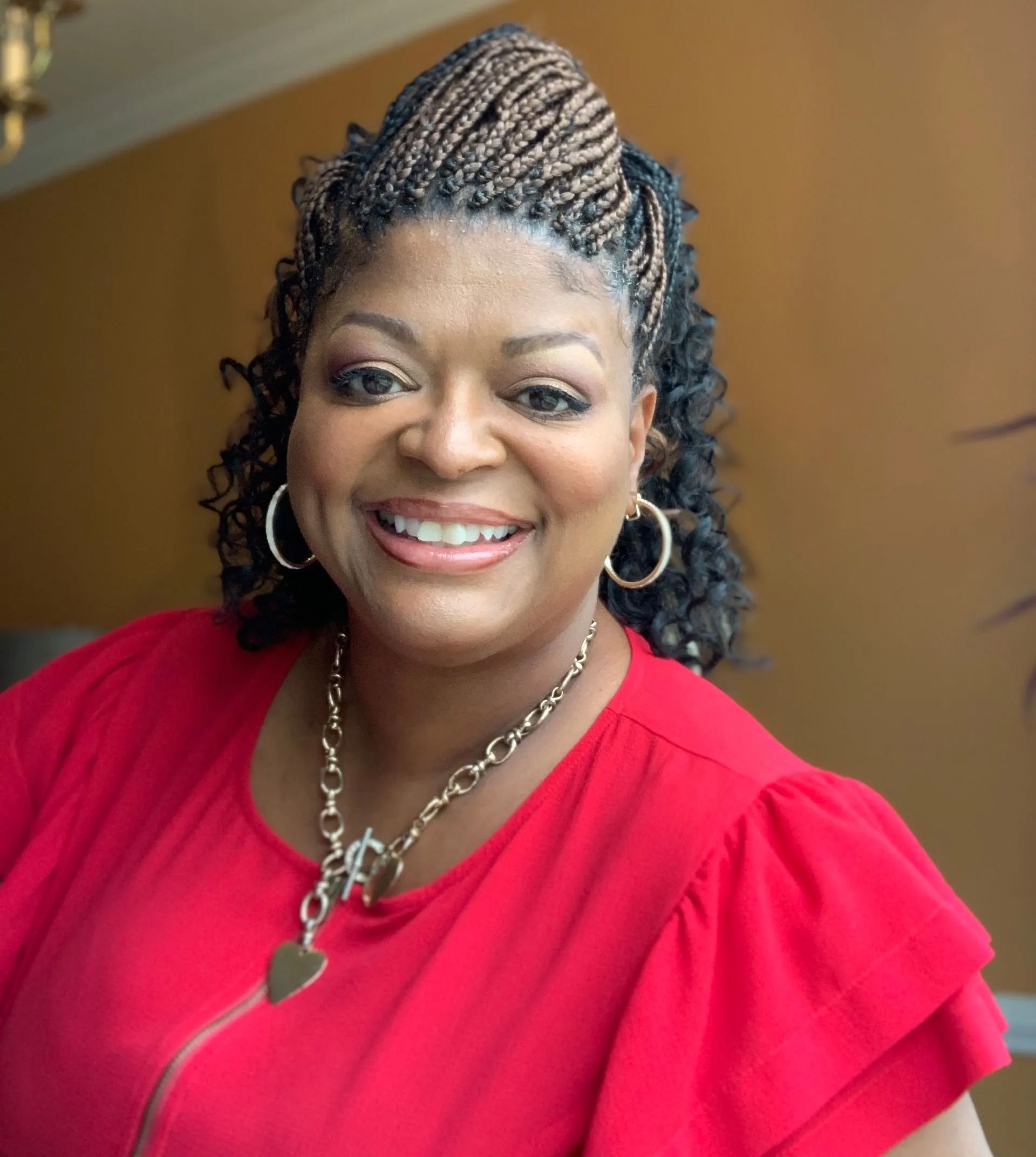 Smiling woman in a red blouse and silver necklace, standing indoors against a warm background