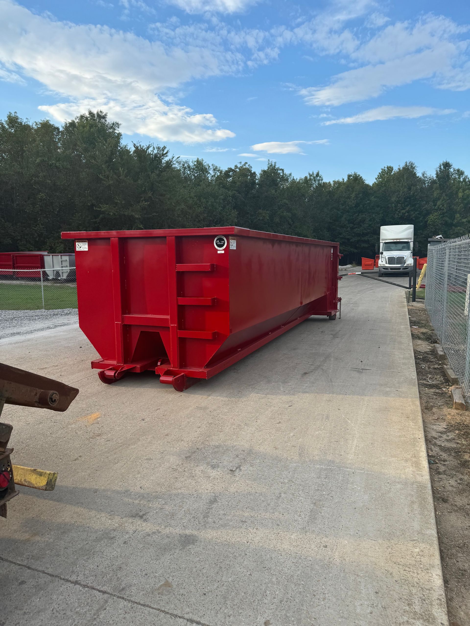 Red construction dumpster container on gravel lot with trees and trucks in the background