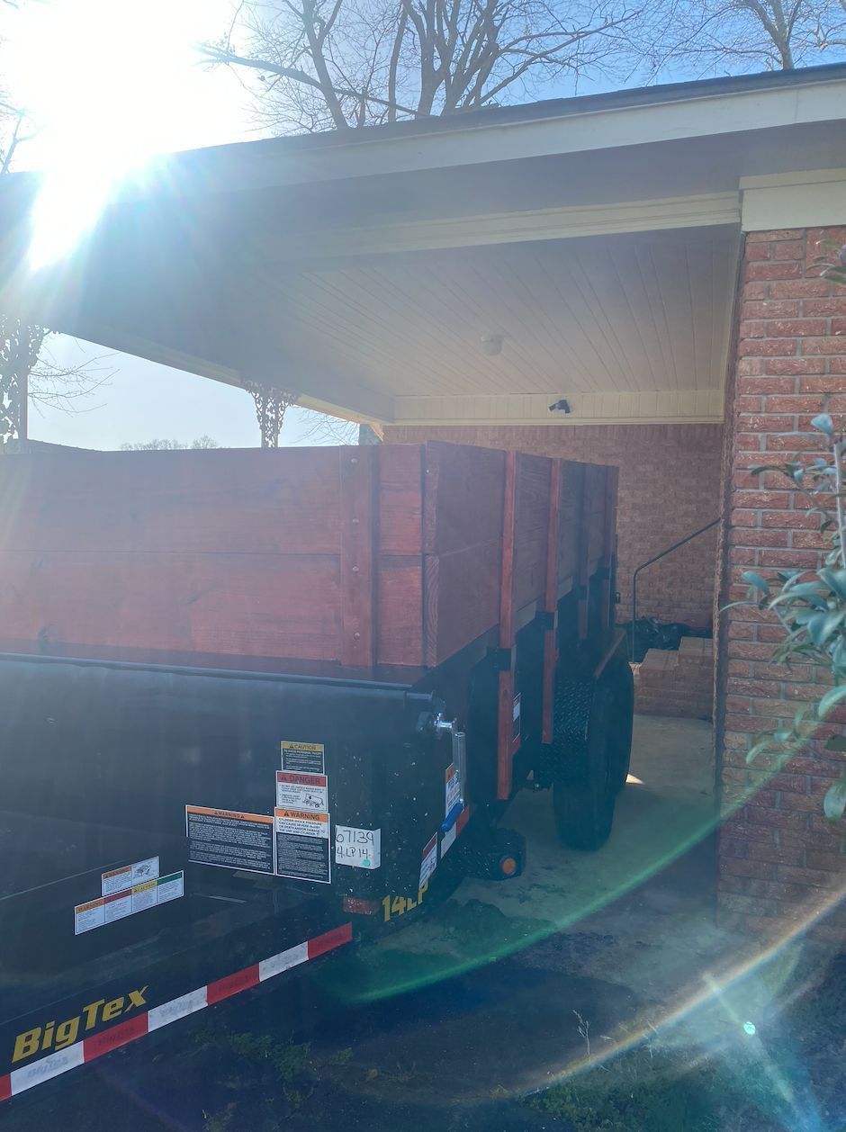 Covered black trailer parked under a carport beside a brick house, with red cargo and sun glare