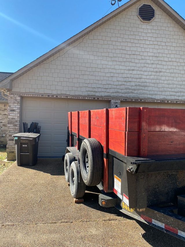 Red dump trailer with black and white wheels parked in a driveway in front of a garage.