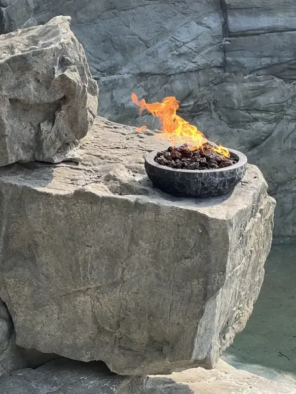 Fire pit with orange flames on a stone, beside a water feature.