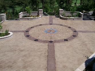 Stone patio with circular design, brown and beige tones, planters with greenery in background.