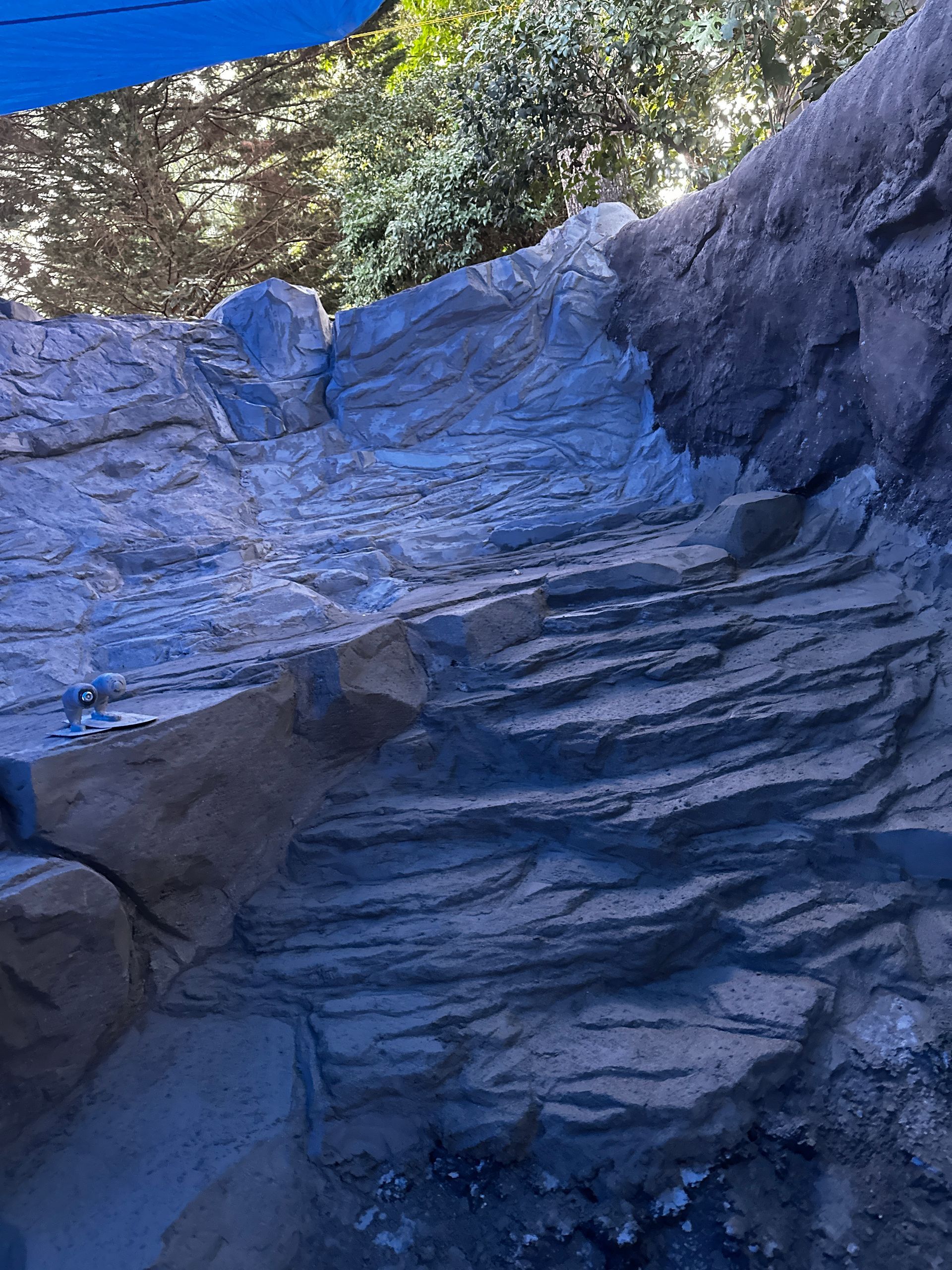 Blue and tan rock wall, possibly artificial, with layered texture; sunlit area above.