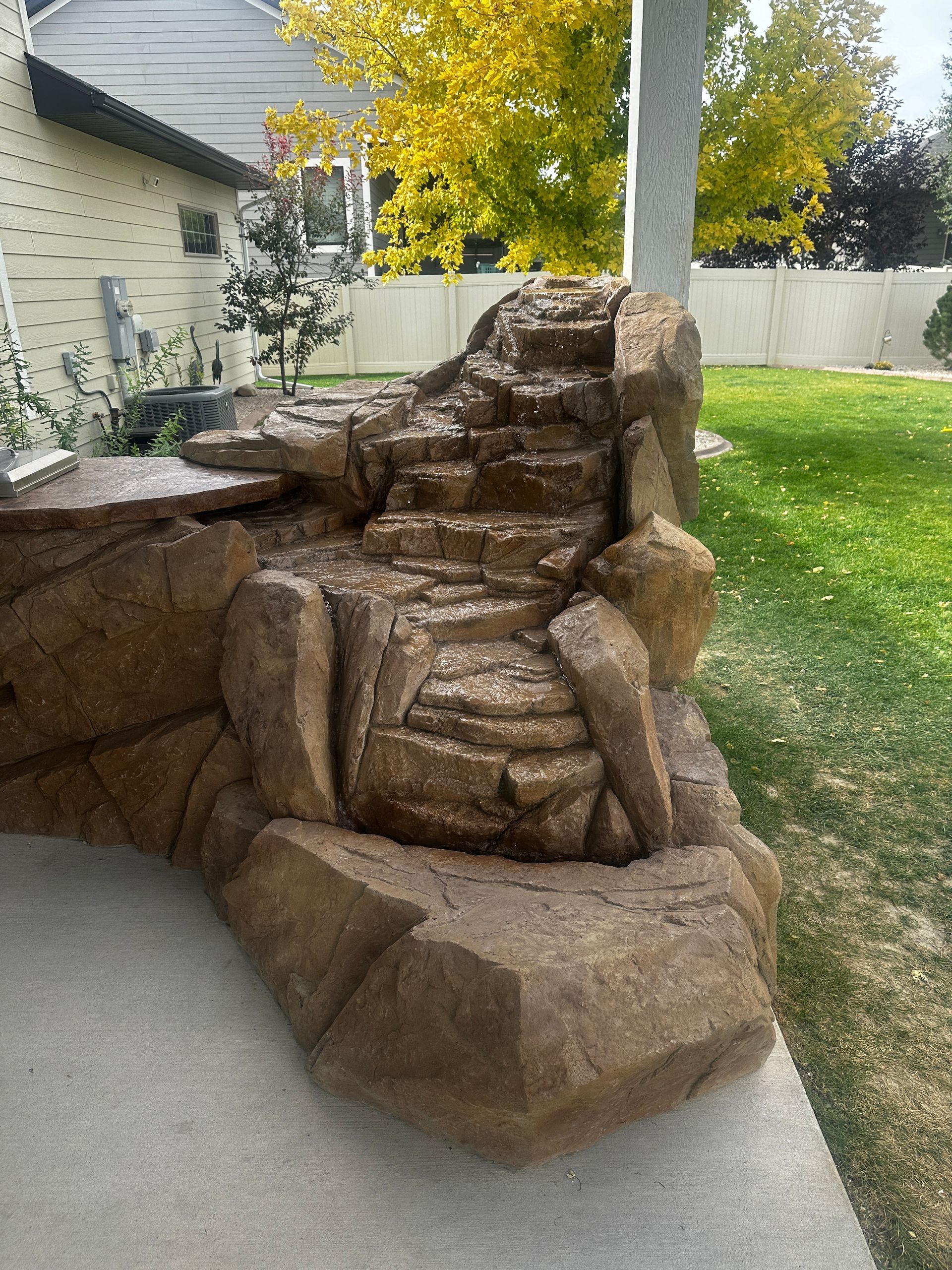 Rock waterfall fountain on a patio with a grassy backyard and trees.