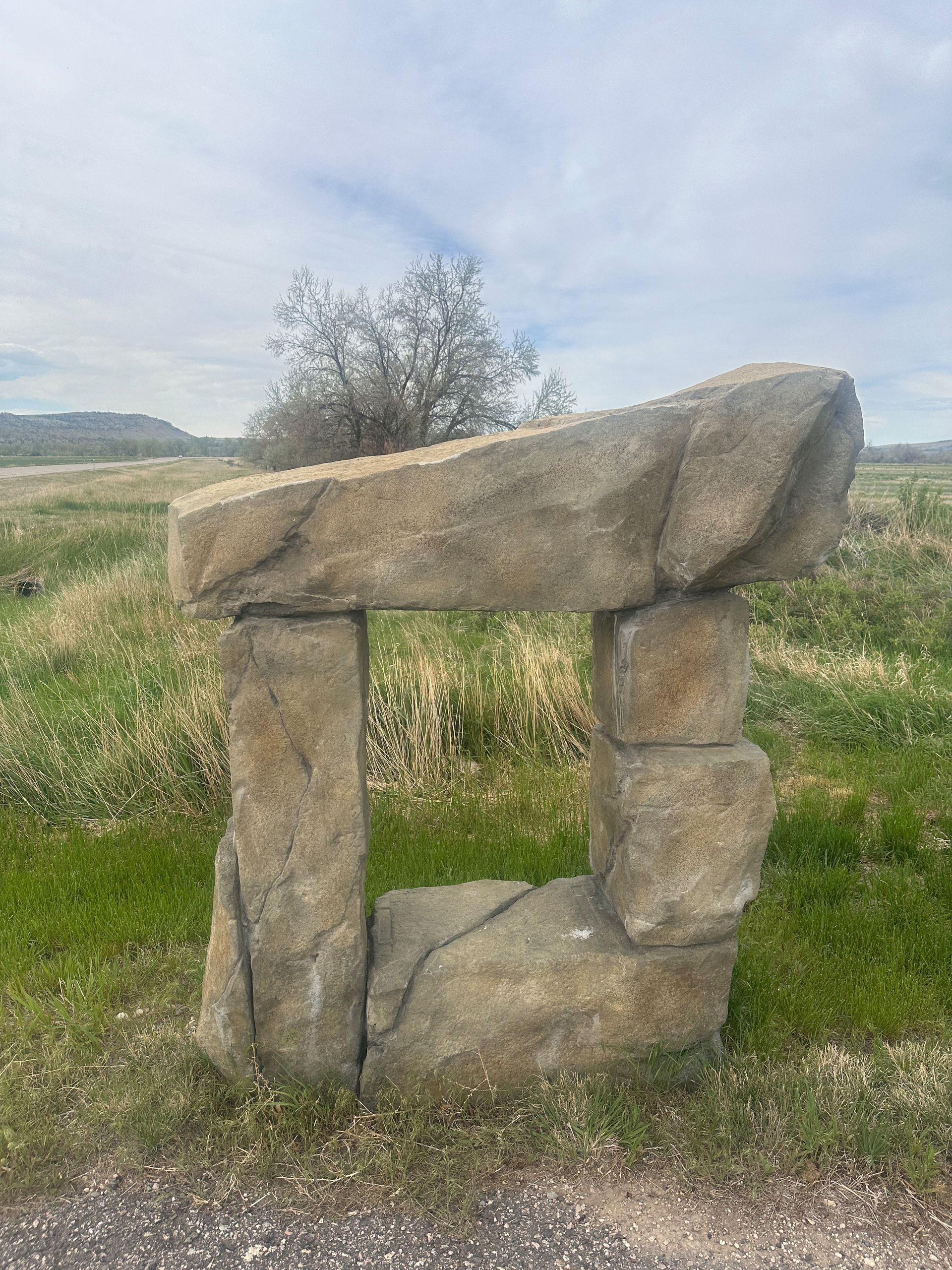 Stone structure in a field, with a square opening. Brownish stones, green grass, and overcast sky.