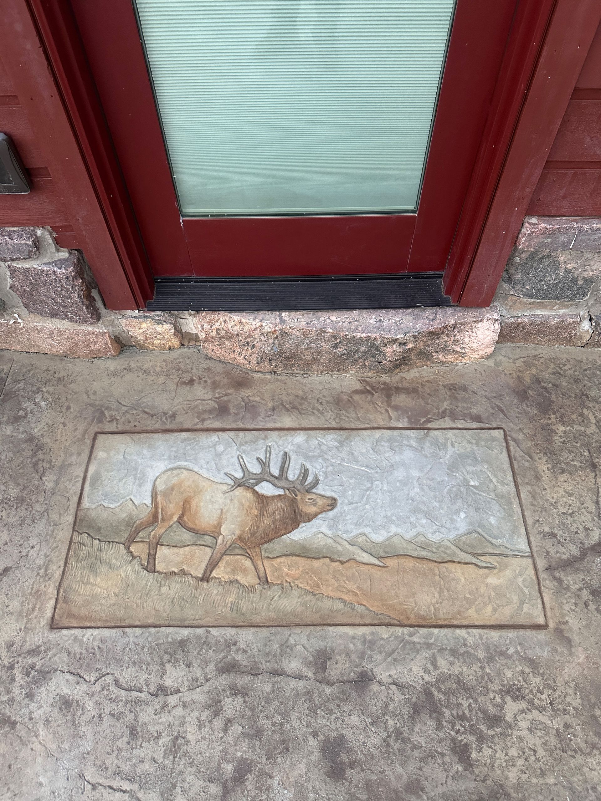Elk art on a rectangular tile set into a stone walkway, in front of a red door with a frosted glass panel.