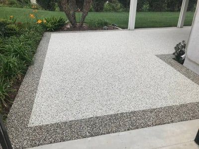 Concrete patio with stone border and light-colored main surface, next to greenery and a white column.