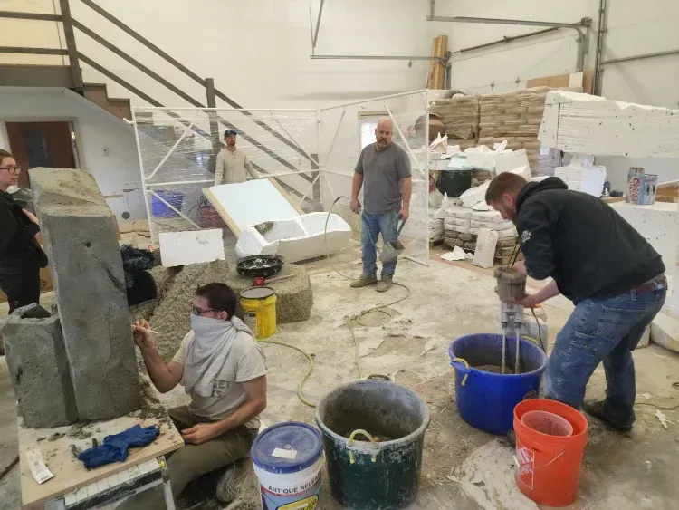 People working in a workshop. One mixes cement, another works on a stone structure, others observe.