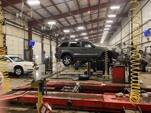 Car on a lift in a repair shop; another car nearby. Tools and equipment visible.