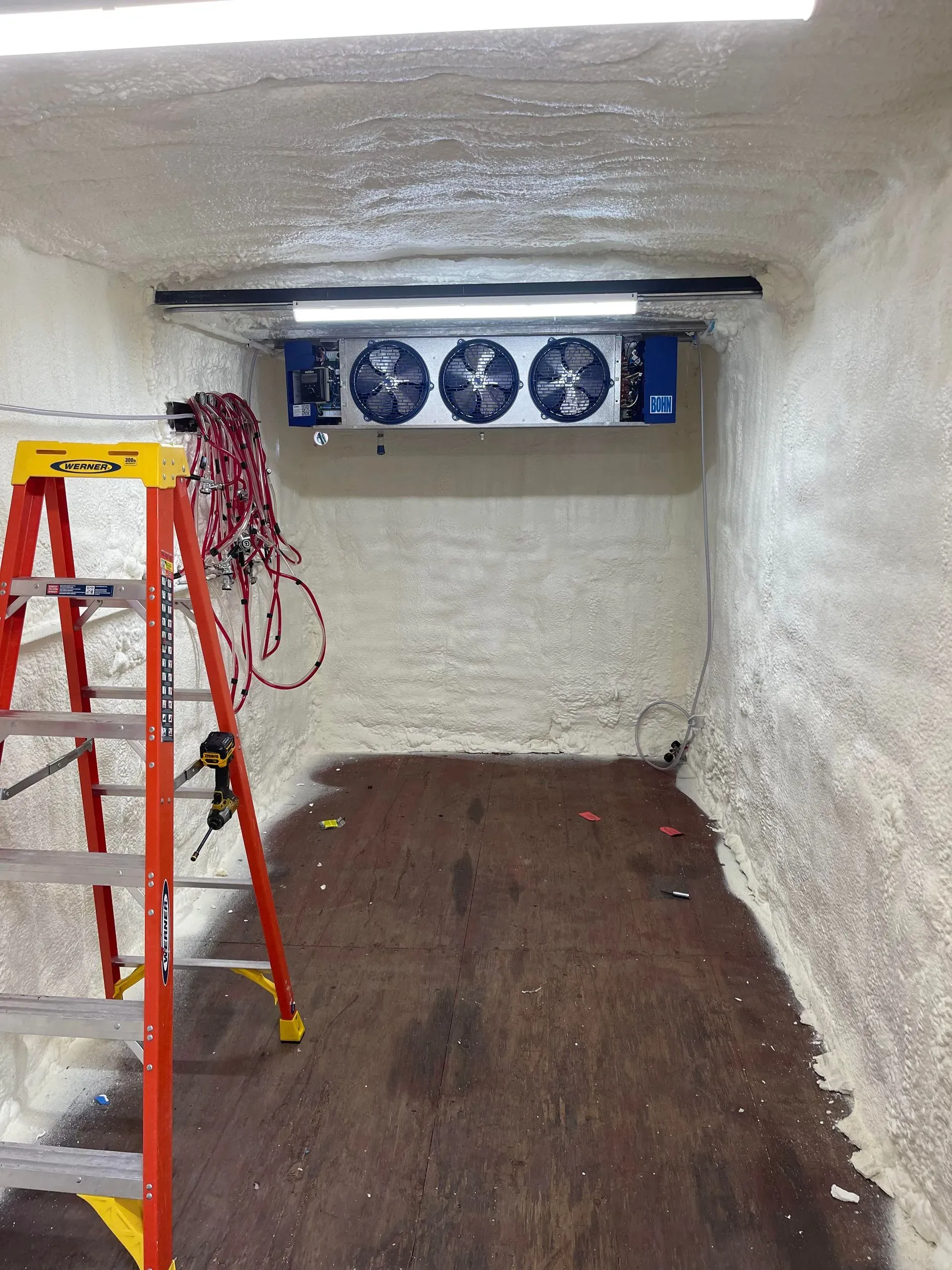 A small cold storage room with a cooling unit, ladder, and wiring. White foam insulation on walls and ceiling.