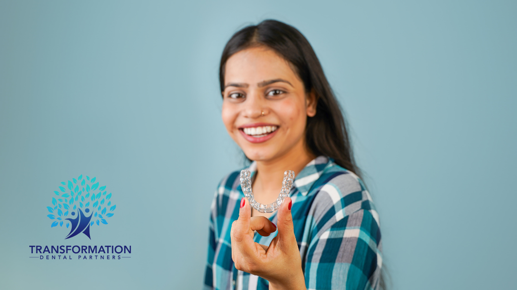 Woman holding clear aligners, smiling at the camera against a blue background, Transformation Dental logo.