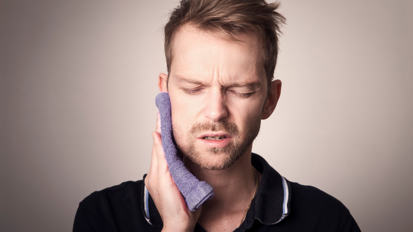 Man holding a cloth to his cheek, wincing in pain against a plain background