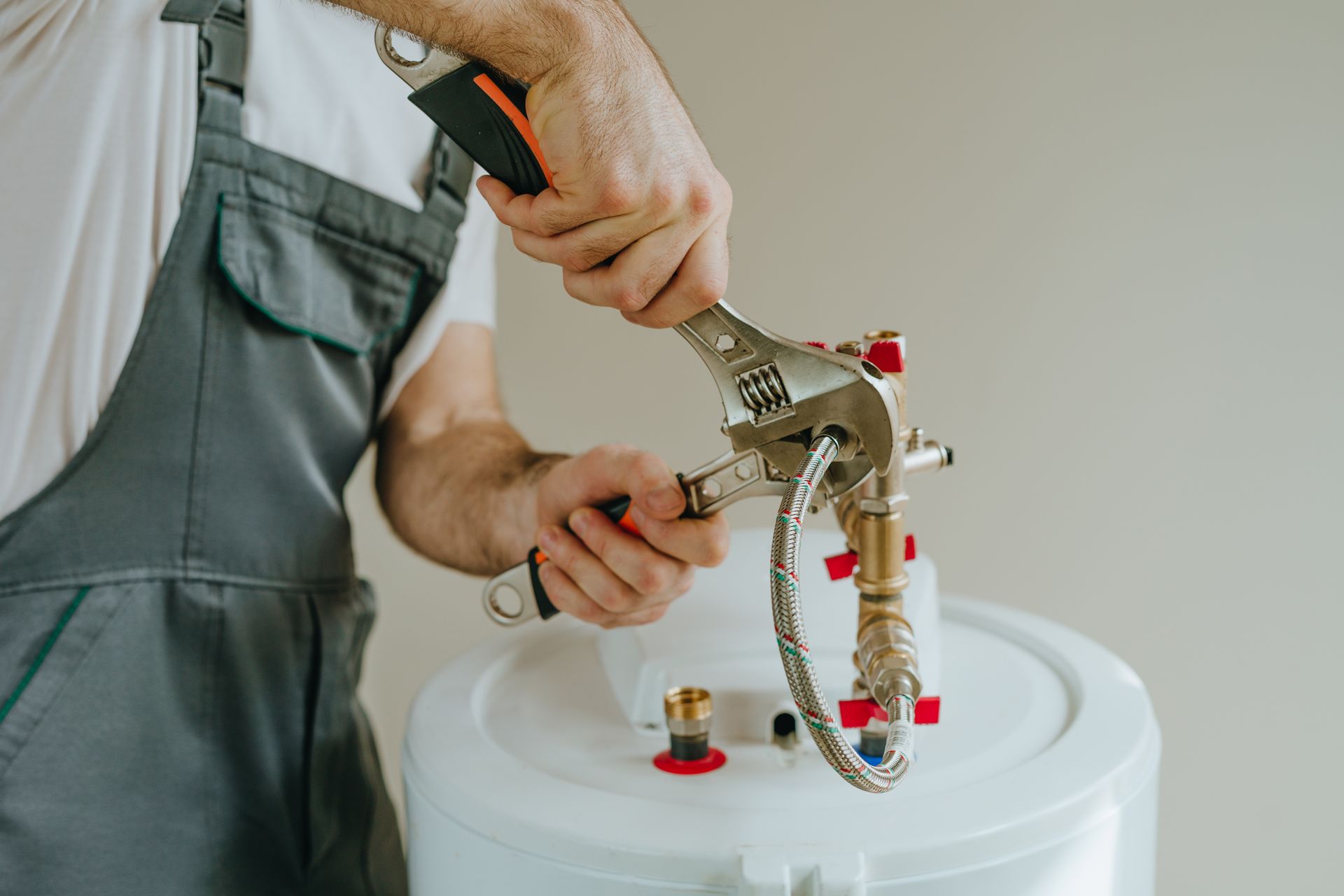 Plumber in gray overalls using a wrench to connect a metal water hose to a white water heater. Plumber in gray overalls using a wrench to connect a metal water hose to a white water heater.