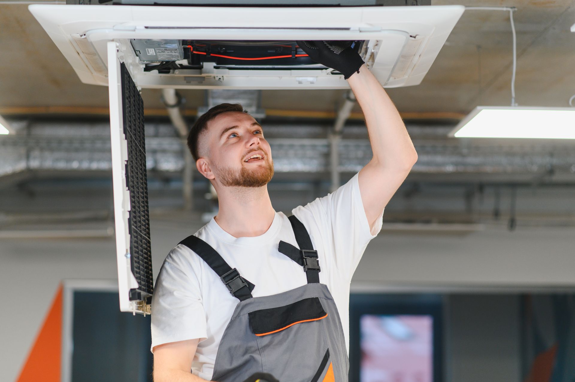 Technician in overalls working on an air conditioning unit in a ceiling, smiling.