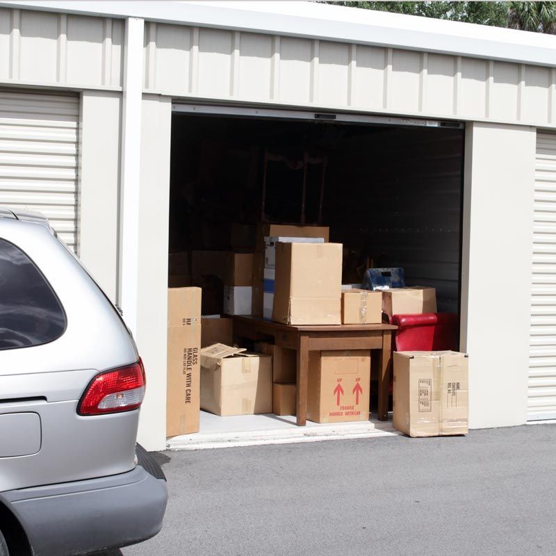 Storage unit filled with stacked cardboard boxes, with a car parked outside the open door.