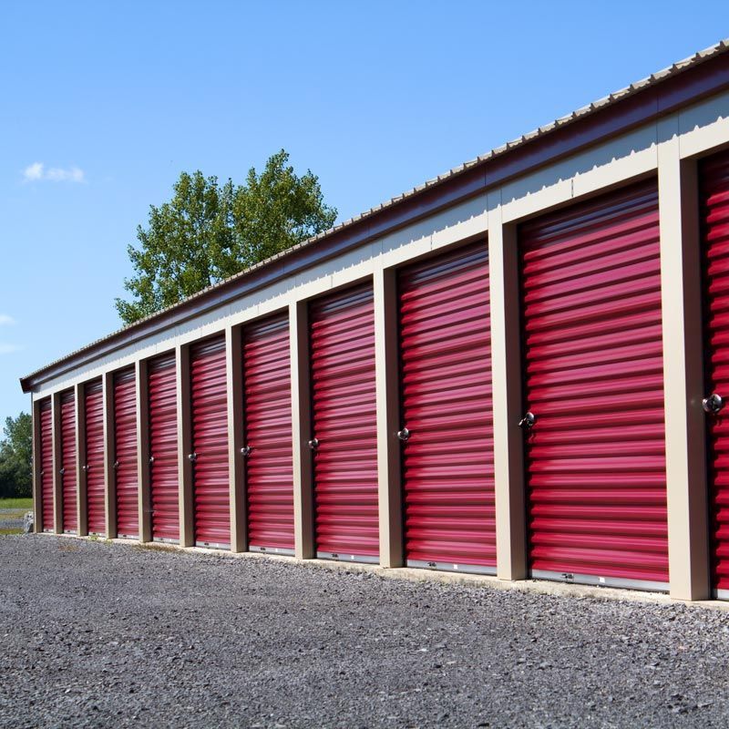 Row of red storage unit doors in a gravel lot under a blue sky
